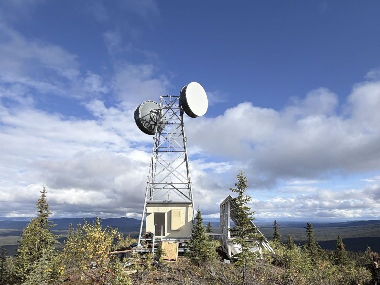Lattice tower with microwave dish antennas and an equipment shelter on a wooded hilltop.